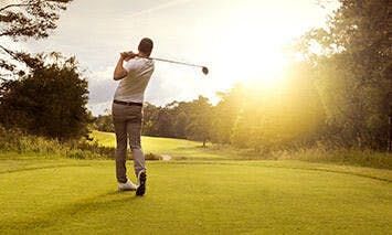A golfer swings a club on a well-maintained green golf course at sunset, surrounded by trees and a clear sky.
