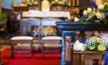 Bouquet, resting, on a wooden pew decorated with white and blue flowers and green leaves. In the background, there are two wooden kneelers on a red carpet in front of an altar.
