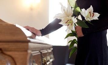 A person dressed in black holds white lilies, standing beside a wooden coffin in a softly lit room.