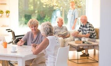 Elderly individuals sit and converse around a bright, homely lounge; two women engage at a table while two men sit on a couch, with a caregiver standing nearby.